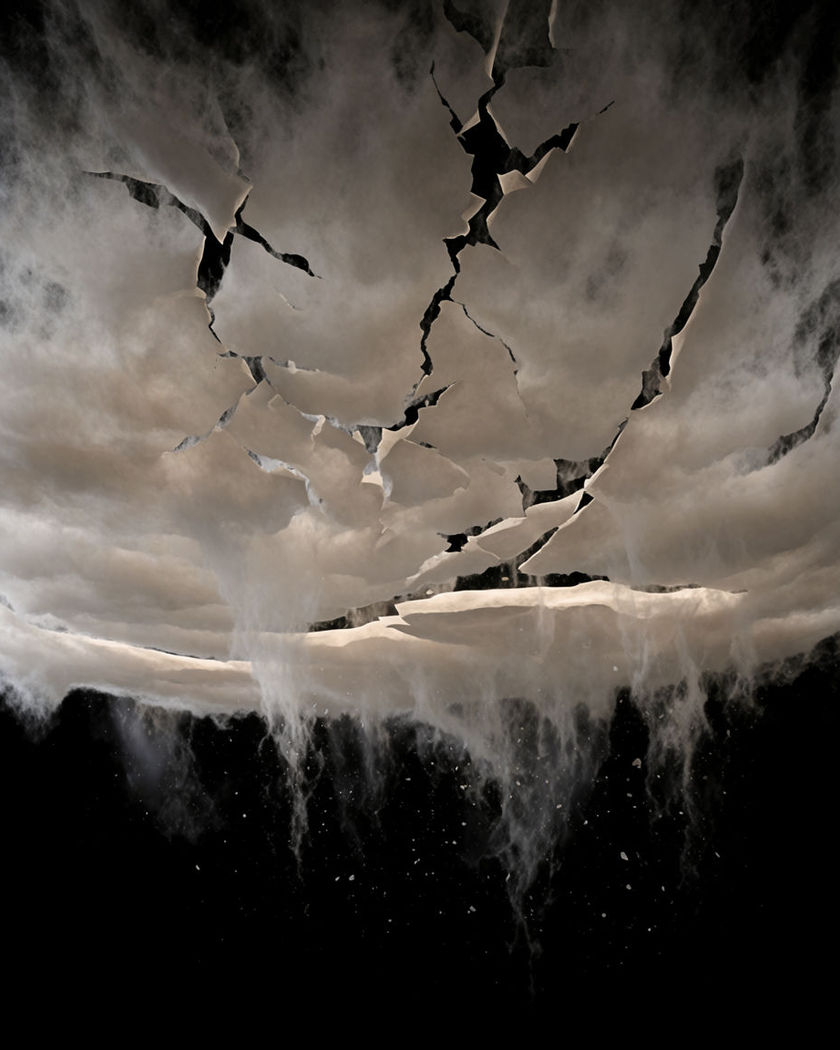 A vast dark ceiling of cloud cracked into descending shards over a pure black lower ground, one thin band of pale light along the seam