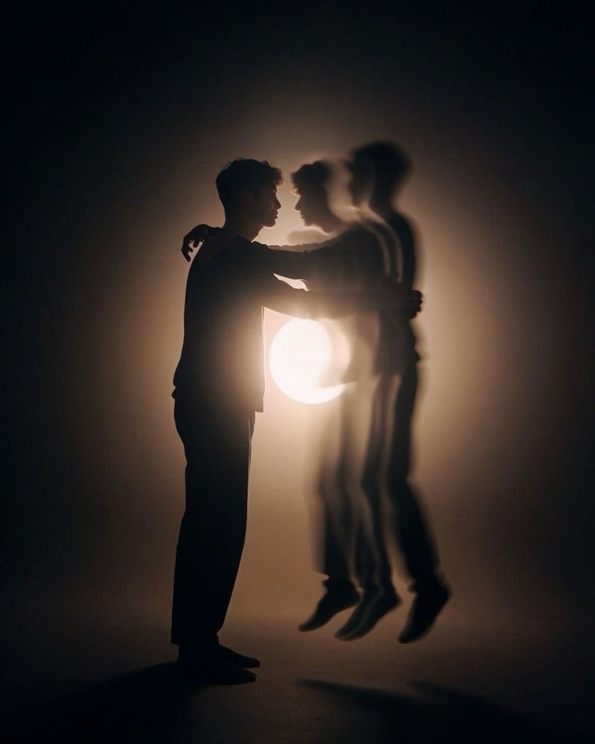 Two silhouetted figures embracing as one figure dissolves and reforms into someone new, soft warm backlight