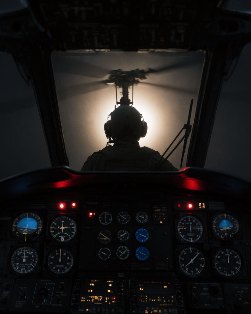 A pilot in a helicopter cockpit, instrument lights glowing as the rotor spins up outside the windshield