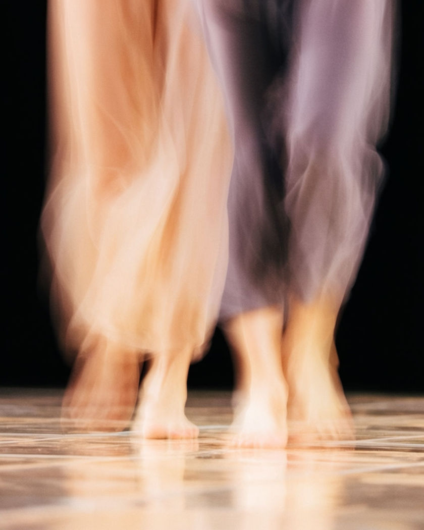 Two blurred streaks of stepping feet caught mid-shuffle over glossy tile, warm rhythmic motion against a pure black ground