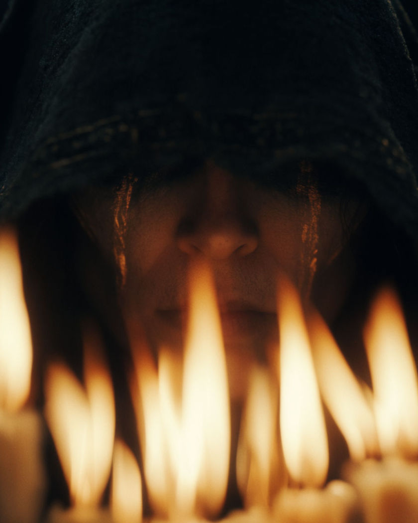 A face with faint glowing ritual markings along the cheekbones, candlelight rising around a dark ceremonial hood