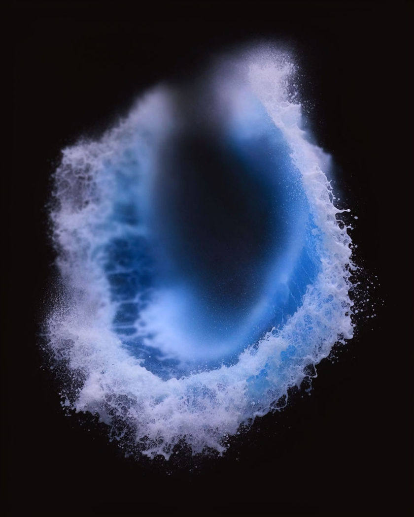 A hem of surf rendered as an abstract ring of foam and spray thrown outward across a on a black ground