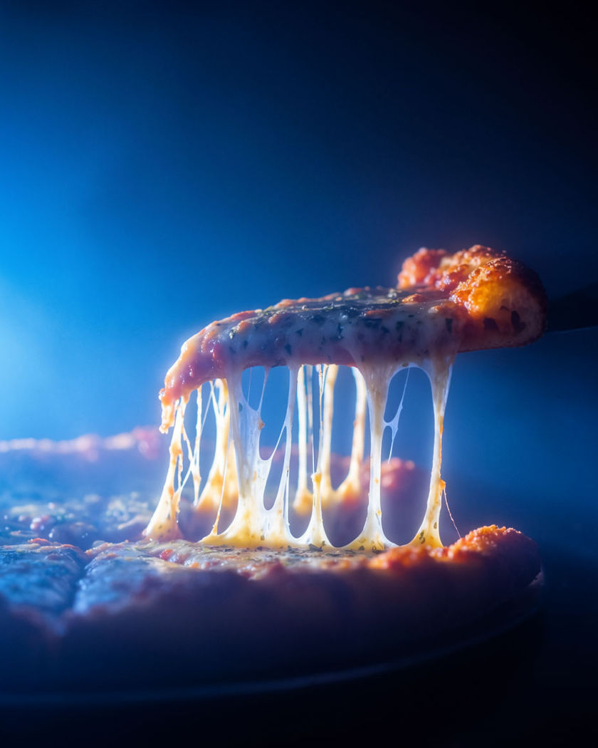 A single slice lifting from a whole pizza with molten cheese stretching into glossy strings against a dark ground