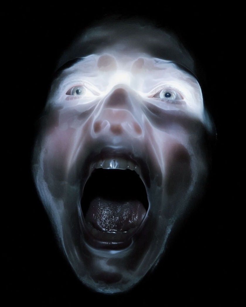 A face mid-scream lit by a single cold light against deep shadow, eyes wide and mouth open