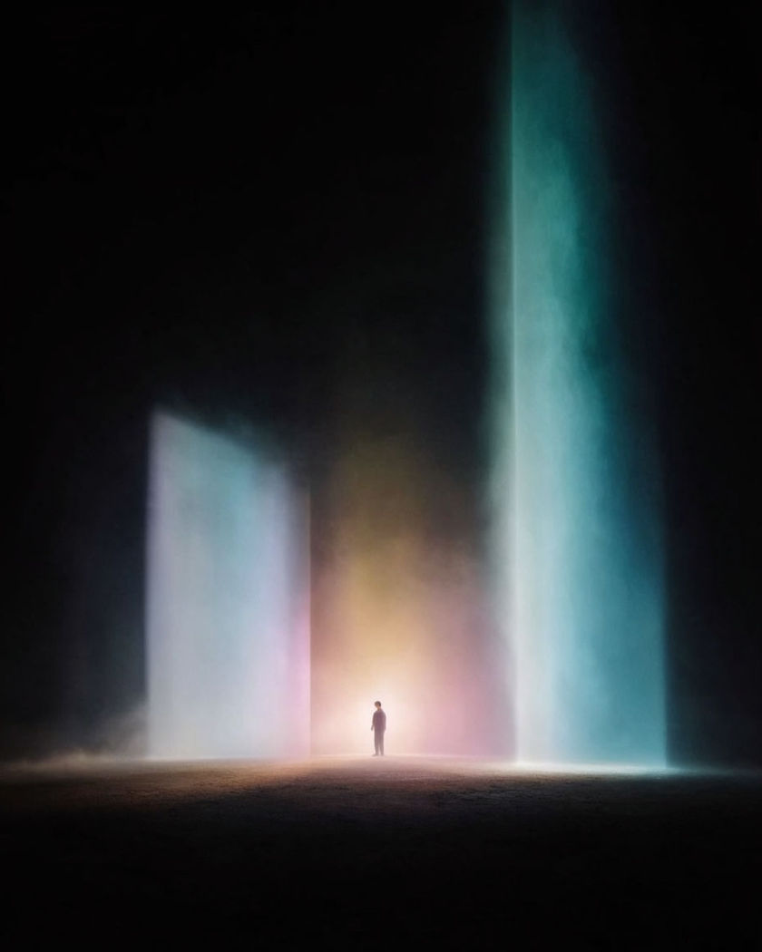 A lone figure spotlit at the centre of a vast dark stage, columns of coloured light and haze rising around them on a black ground
