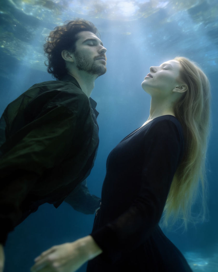 Two people floating together underwater in soft blue light with fabric drifting around them