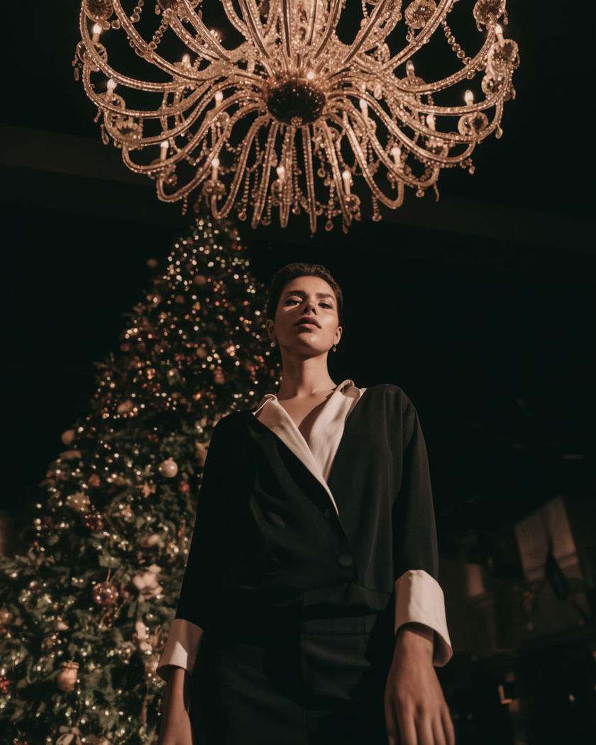 A person in elegant evening wear standing beneath a grand chandelier at a Christmas gala with a decorated tree glowing behind